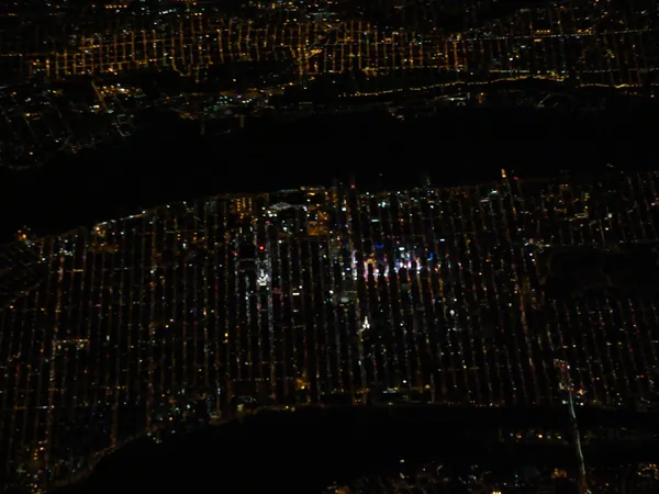 Manhattan at night, with Times Square glowing brightest and the perfectly straight street grid clearly visible across the island. At the top, the dark band is the Hudson River.
